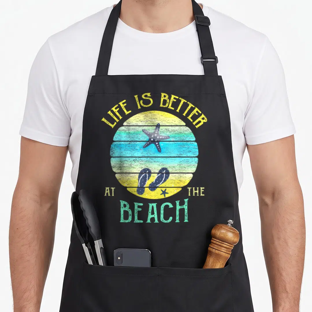 Better beach life apron - Life is Better at the Beach text over striped sunset with starfish and flip flops — shown on a black cotton canvas apron against a white background