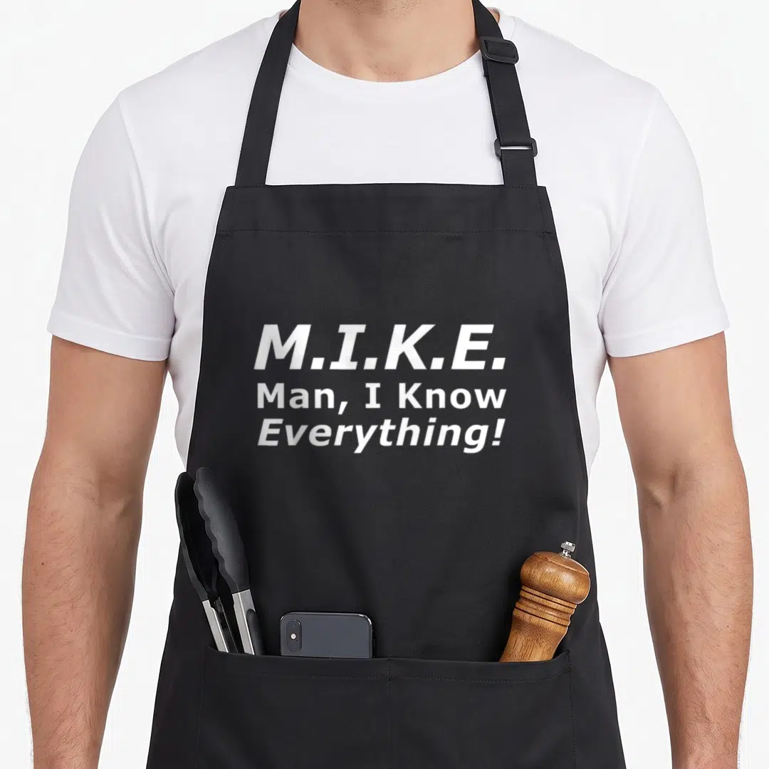 Mike e.a apron - White stacked text M.I.K.E. Man I Know Everything on dark background — shown on a black cotton canvas apron against a white background
