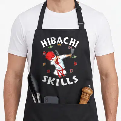 Connoisseur hibachi apron - Cartoon chef holding knife and cleaver surrounded by sushi with HIBACHI SKILLS text — shown on a black cotton canvas apron against a white background