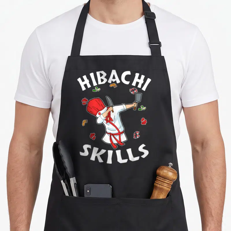 Connoisseur hibachi apron - Cartoon chef holding knife and cleaver surrounded by sushi with HIBACHI SKILLS text — shown on a black cotton canvas apron against a white background