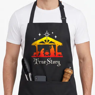 Story jesus nativity christmas apron - Nativity silhouette under stable with stars and True Story text in yellow orange — shown on a black cotton canvas apron against a white background