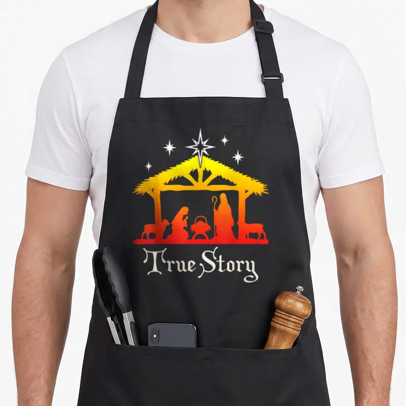 Story jesus nativity christmas apron - Nativity silhouette under stable with stars and True Story text in yellow orange — shown on a black cotton canvas apron against a white background
