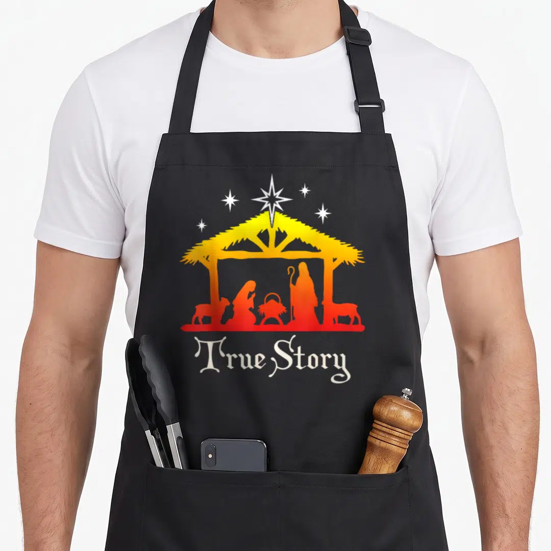 Story jesus nativity christmas apron - Nativity silhouette under stable with stars and True Story text in yellow orange — shown on a black cotton canvas apron against a white background