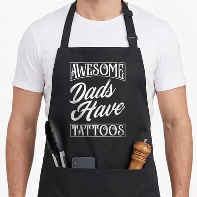 Tattooed father tattoo aprons - White stacked typography reading AWESOME DADS HAVE TATTOOS with framed border elements — shown on a black cotton canvas apron against a white background