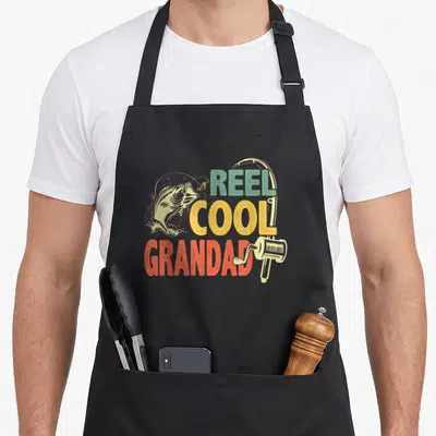 Reel grandad aprons - Retro REEL COOL GRANDAD text with jumping fish and fishing reel in teal yellow red beige — shown on a black cotton canvas apron against a white background