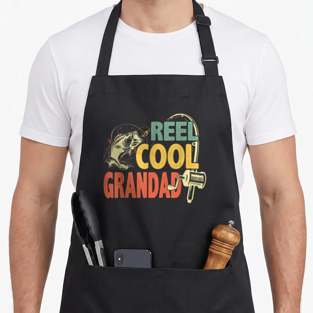 Reel grandad aprons - Retro REEL COOL GRANDAD text with jumping fish and fishing reel in teal yellow red beige — shown on a black cotton canvas apron against a white background