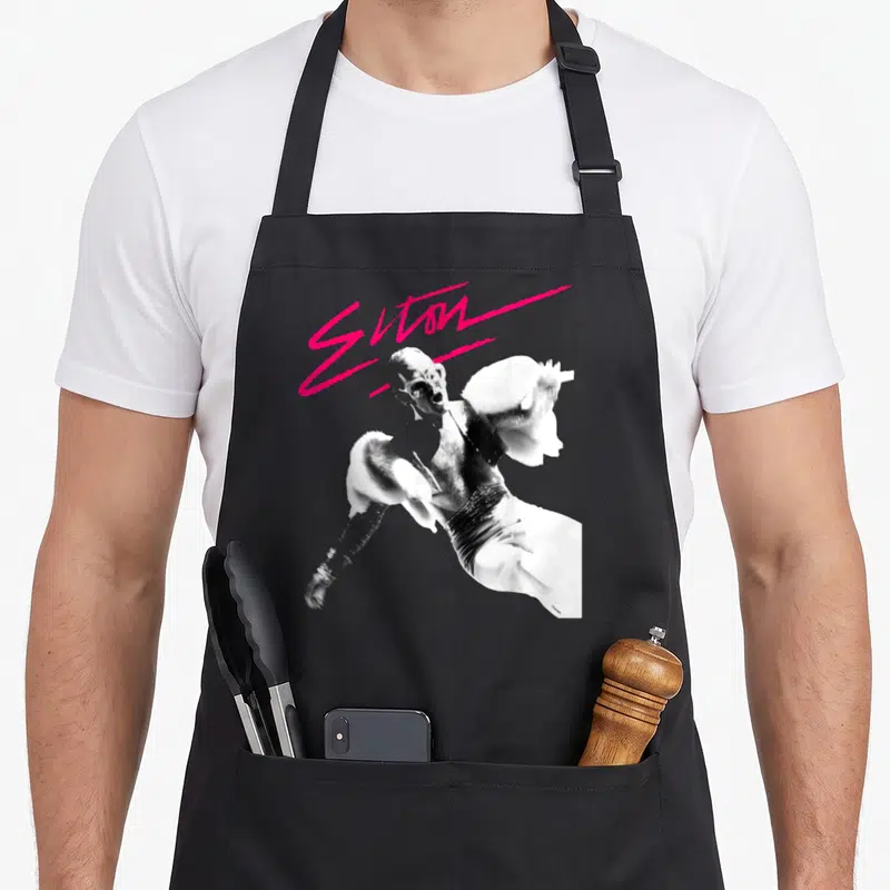 Elton birthday pink apron - Pink script reading Elton above black and white photo of man singing — shown on a black cotton canvas apron against a white background