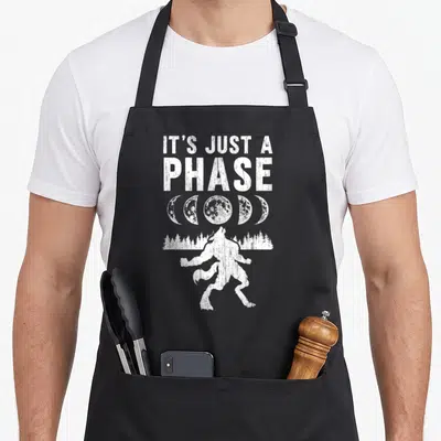 Phase werewolf apron - White distressed IT'S JUST A PHASE text, five moon phases, and a howling werewolf silhouette — shown on a black cotton canvas apron against a white background