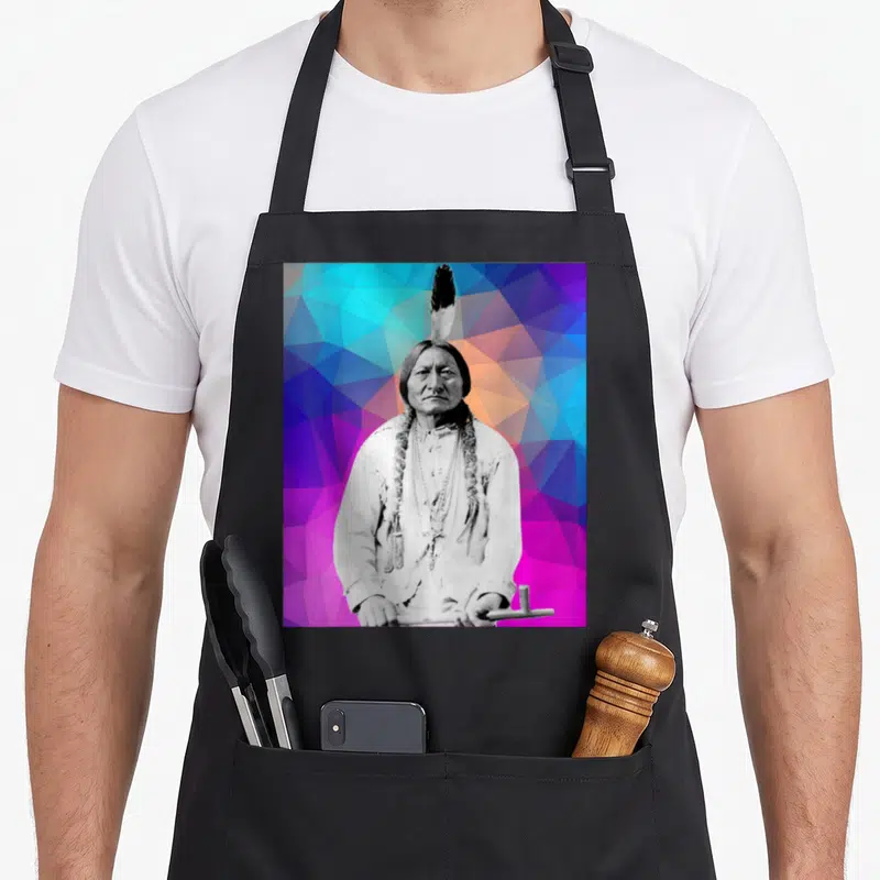 Bull native american apron - Grayscale Sitting Bull portrait holding pipe against blue purple pink orange geometric — shown on a black cotton canvas apron against a white background