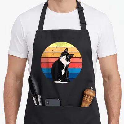 Animal tuxedo apron - Black and white cat sitting in front of yellow orange red and blue horizontal stripes — shown on a black cotton canvas apron against a white background