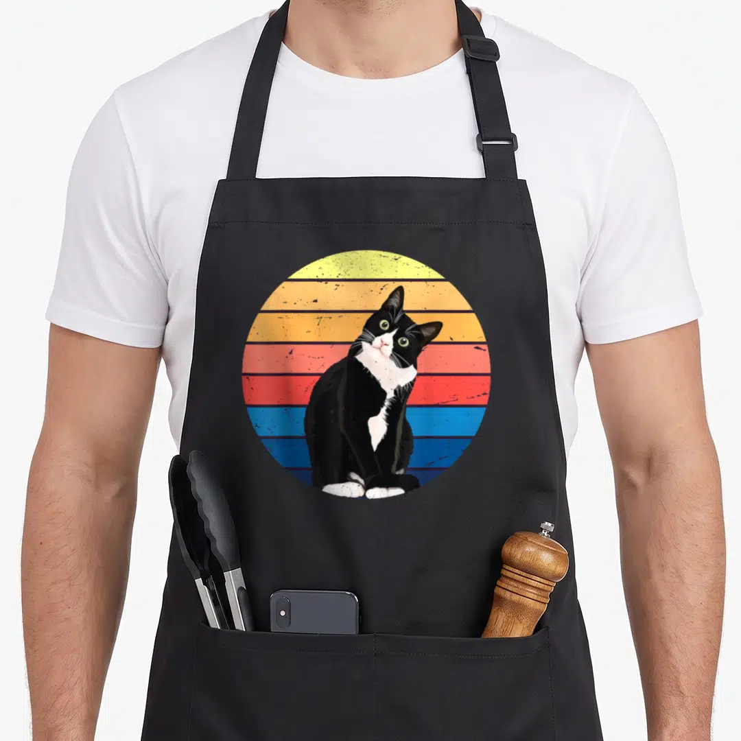Animal tuxedo apron - Black and white cat sitting in front of yellow orange red and blue horizontal stripes — shown on a black cotton canvas apron against a white background