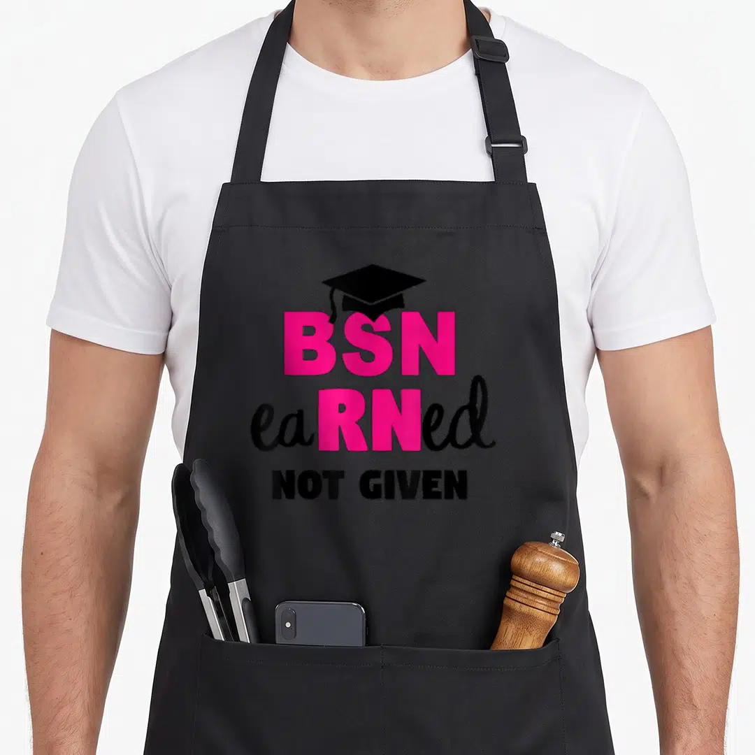 Earned nursing aprons - BSN earned not given text with black graduation cap in pink and black typography — shown on a black cotton canvas apron against a white background