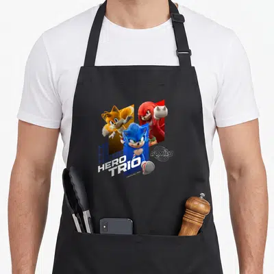 Hero hedgehog owner apron - Three cartoon hedgehogs and fox over blue red yellow blocks with stacked HERO TRIO text — shown on a black cotton canvas apron against a white background