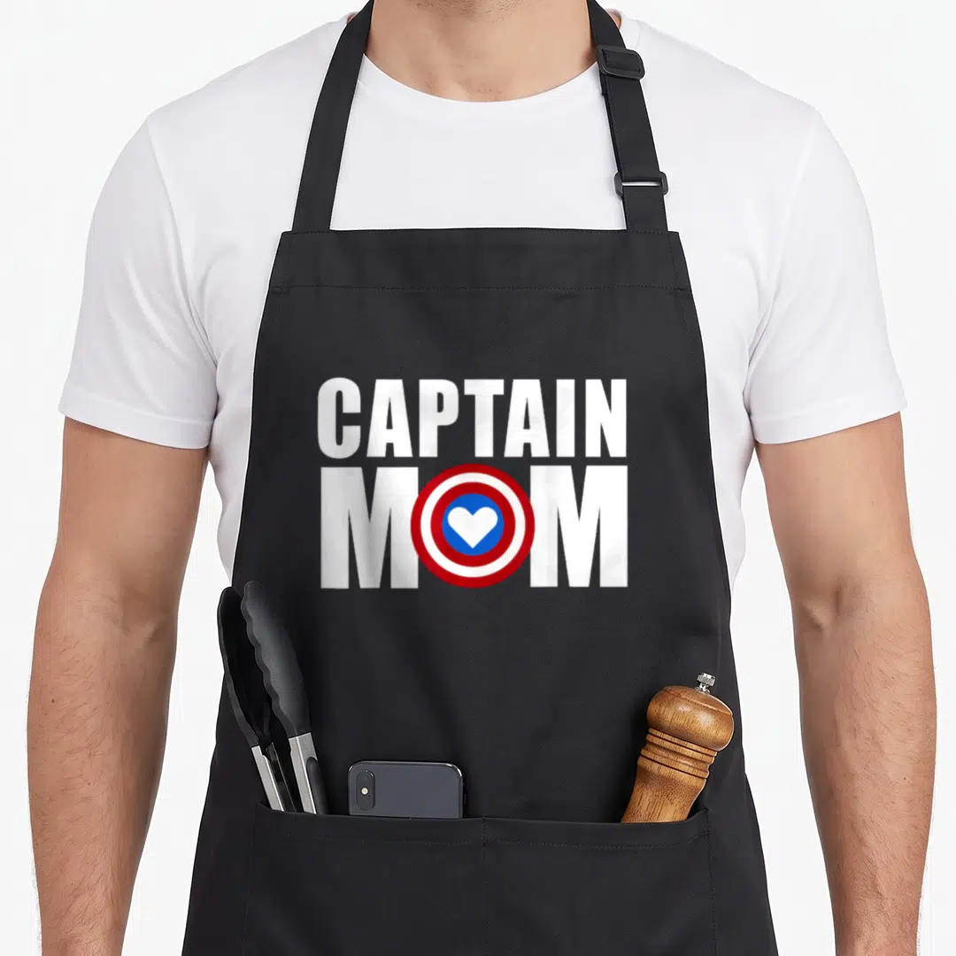 Captain mother's apron - CAPTAIN MOM text with red white blue shield heart replacing the O — shown on a black cotton canvas apron against a white background