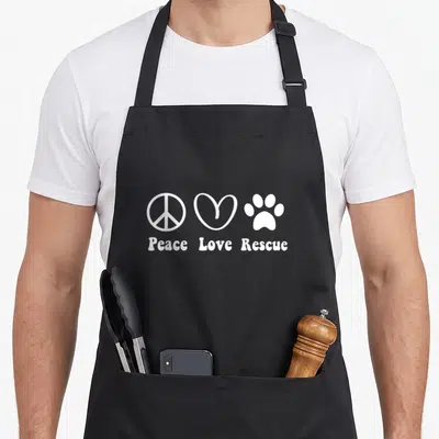 Peace grill rescue apron - White peace sign, heart, and paw print with text Peace Love Rescue — shown on a black cotton canvas apron against a white background