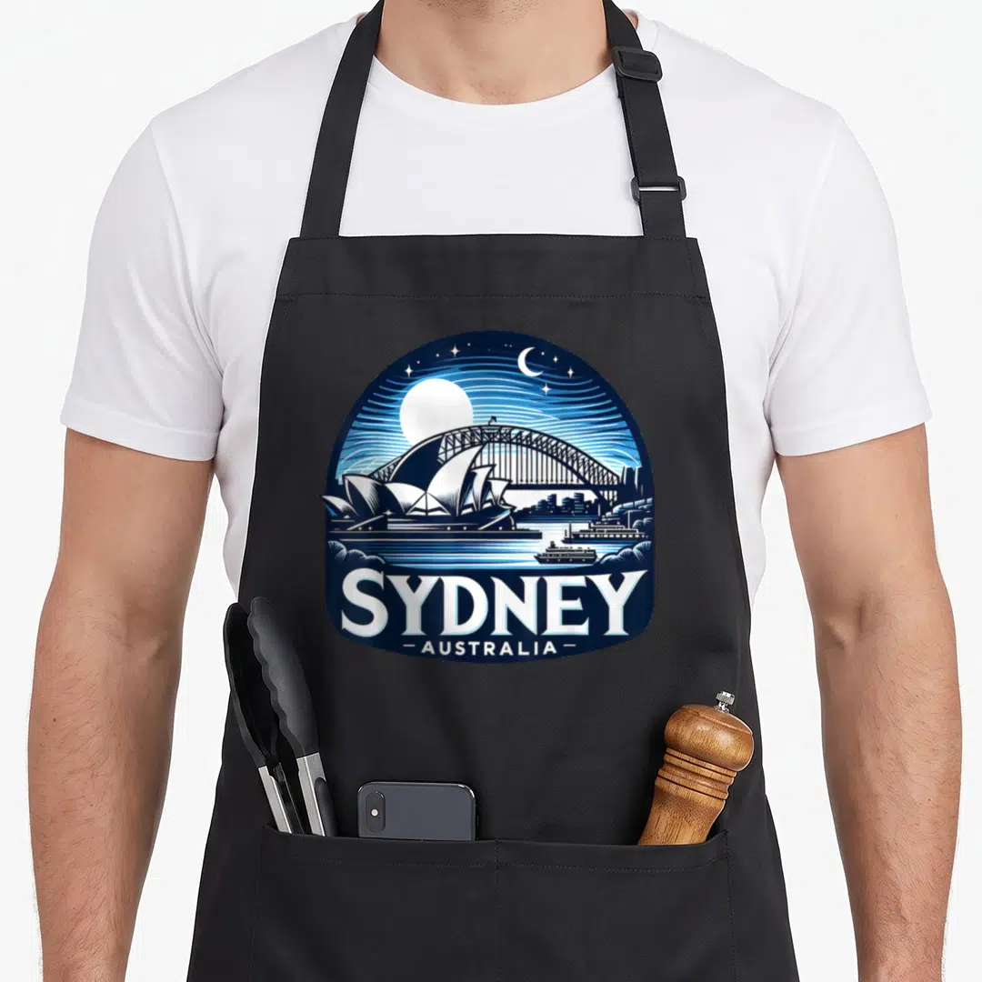 Sydney opera apron - Circular navy blue night skyline print with Opera House, Harbour Bridge, moon, and SYDNEY — shown on a black cotton canvas apron against a white background
