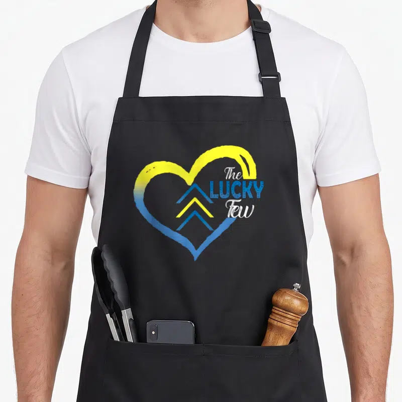Syndrome tattoo apron - Yellow and blue heart with three chevrons and text The Lucky Few — shown on a black cotton canvas apron against a white background