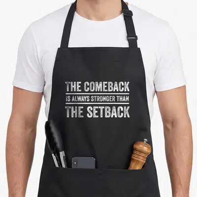 Stronger comeback apron - White distressed text reading THE COMEBACK IS ALWAYS STRONGER THAN THE SETBACK separated — shown on a black cotton canvas apron against a white background