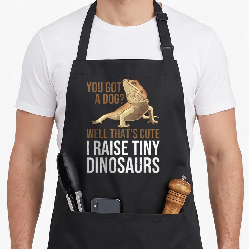 Bearded tiny chef apron - Realistic bearded dragon with You Got A Dog text and I RAISE TINY DINOSAURS slogan in tan — shown on a black cotton canvas apron against a white background