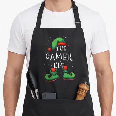 Pajamas christmas elf apron - Stacked text THE GAMER ELF between green hat and pair of shoes with scattered stars — shown on a black cotton canvas apron against a white background