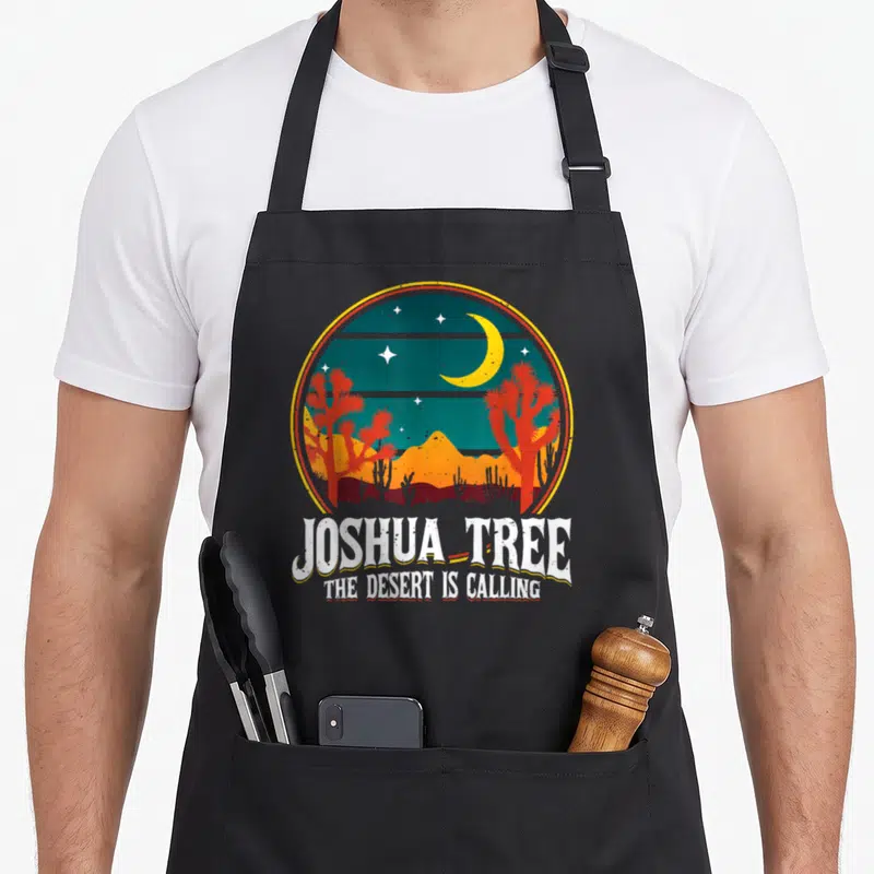 Funny national tree apron - Retro circular desert scene with moon, red trees, and white JOSHUA TREE text — shown on a black cotton canvas apron against a white background