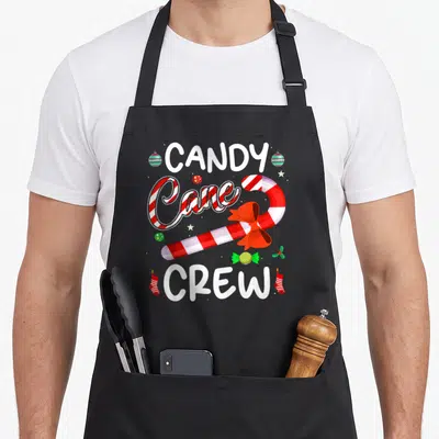Crew candy cane apron - CANDY CREW text surrounding a red bow candy cane with green ornaments and stockings — shown on a black cotton canvas apron against a white background