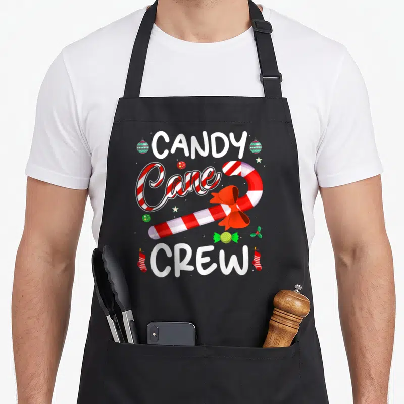 Crew candy cane apron - CANDY CREW text surrounding a red bow candy cane with green ornaments and stockings — shown on a black cotton canvas apron against a white background