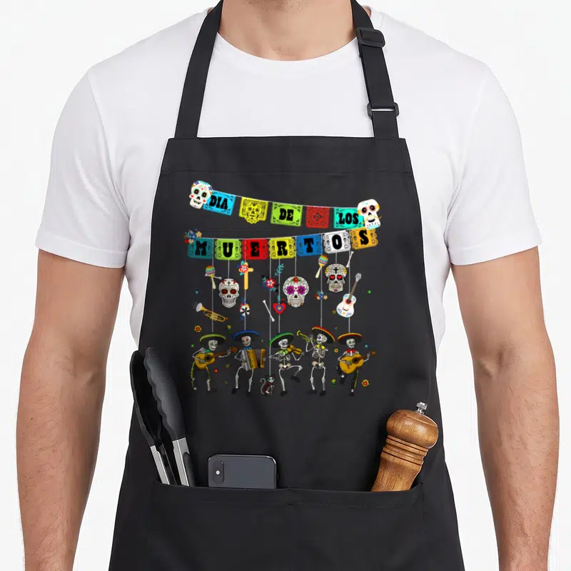 Dancing day of the dead apron - Skeleton mariachi band under Dia De Los Muertos banners with hanging skulls and cat — shown on a black cotton canvas apron against a white background