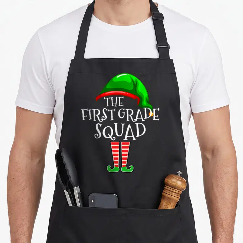 Grade christmas aprons - Cartoon elf hat and shoes framing The First Grade Squad text in green red and white — shown on a black cotton canvas apron against a white background