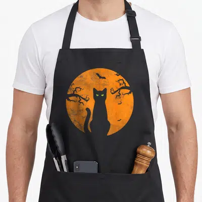 Christmas black cat apron - Grey cat silhouette with green eyes in front of orange striped moon and bats — shown on a black cotton canvas apron against a white background