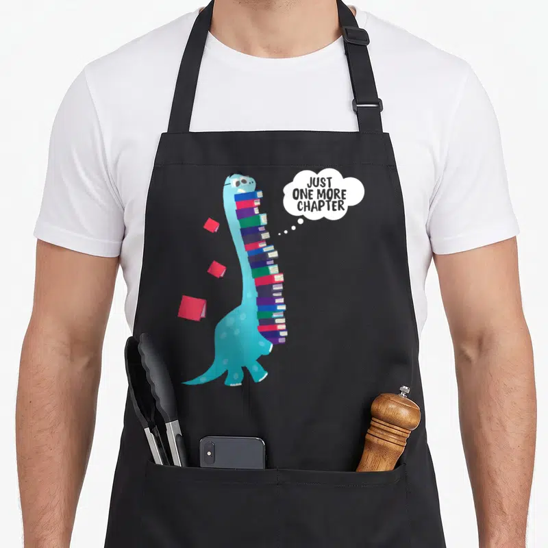 Chapter dinosaur apron - Blue dinosaur holding books with thought bubble JUST ONE MORE CHAPTER — shown on a black cotton canvas apron against a white background