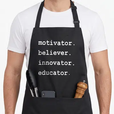 Motivator teacher apron - Four lines of white text reading motivator believer innovator educator — shown on a black cotton canvas apron against a white background