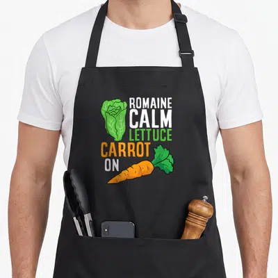 Lettuce romaine calm and carrot on apron - Cartoon green lettuce and orange carrot with white and green text reading — shown on a black cotton canvas apron against a white background