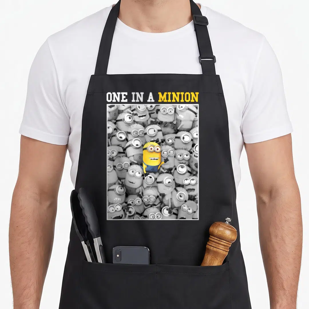 Pop novelty apron - One in a Minion text above yellow minion surrounded by grey minions in white bordered square — shown on a black cotton canvas apron against a white background