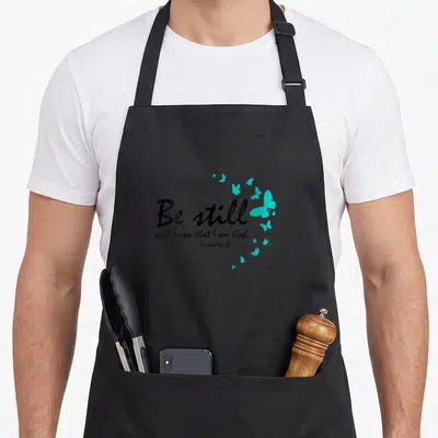 Christian butterfly apron - Black script Be still text with turquoise butterfly cluster on grey background — shown on a black cotton canvas apron against a white background