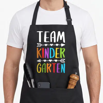 School cute teacher aprons - Stacked TEAM KINDER GARTEN text in rainbow colors separated by white arrows with hearts — shown on a black cotton canvas apron against a white background