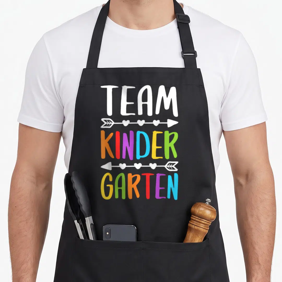 School cute teacher aprons - Stacked TEAM KINDER GARTEN text in rainbow colors separated by white arrows with hearts — shown on a black cotton canvas apron against a white background