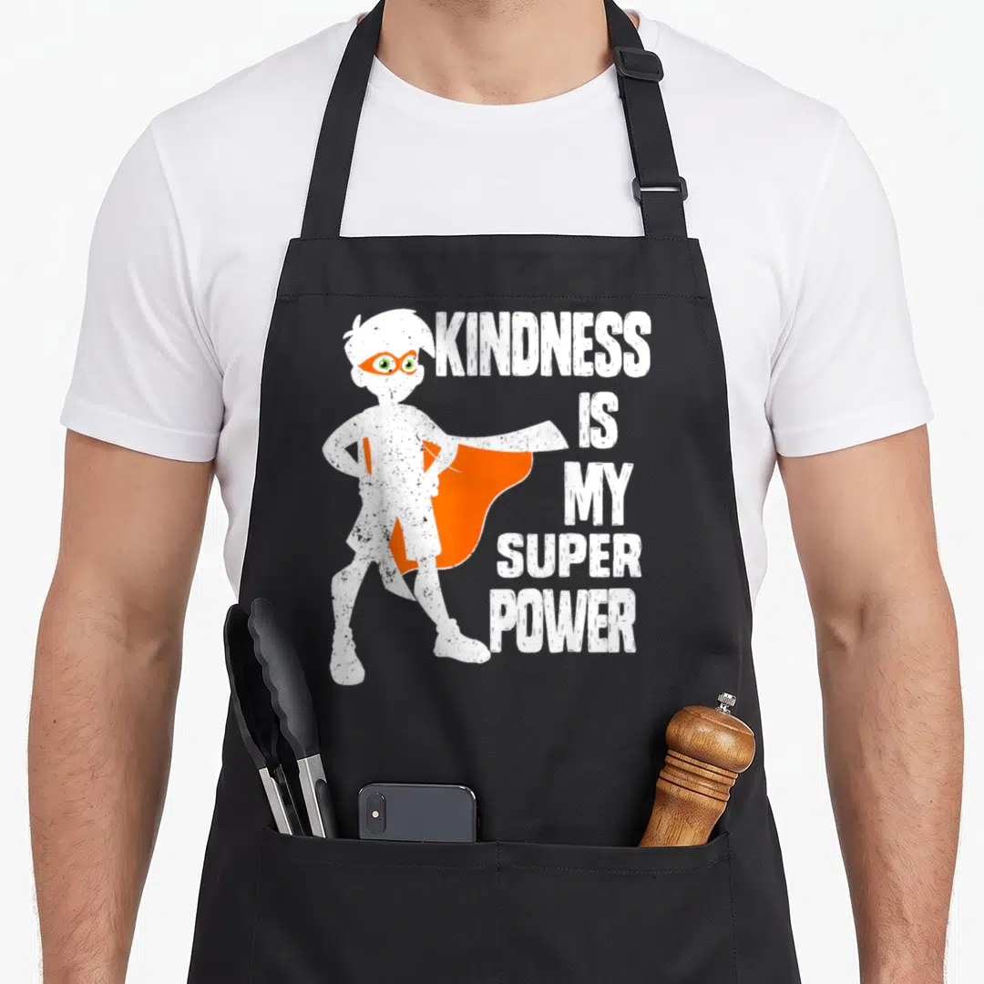 Bullying novelty apron - Cartoon superhero boy with orange cape stands beside stacked white text reading kindness is my — shown on a black cotton canvas apron against a white background