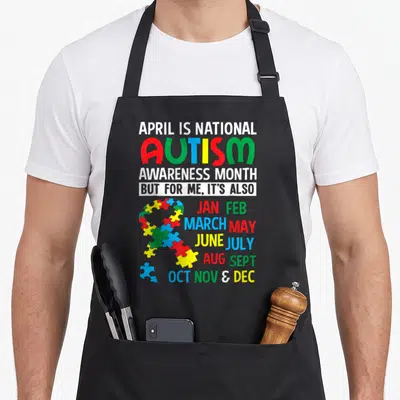 National april apron - Multi-colored puzzle ribbon with stacked text listing all twelve months of the year — shown on a black cotton canvas apron against a white background