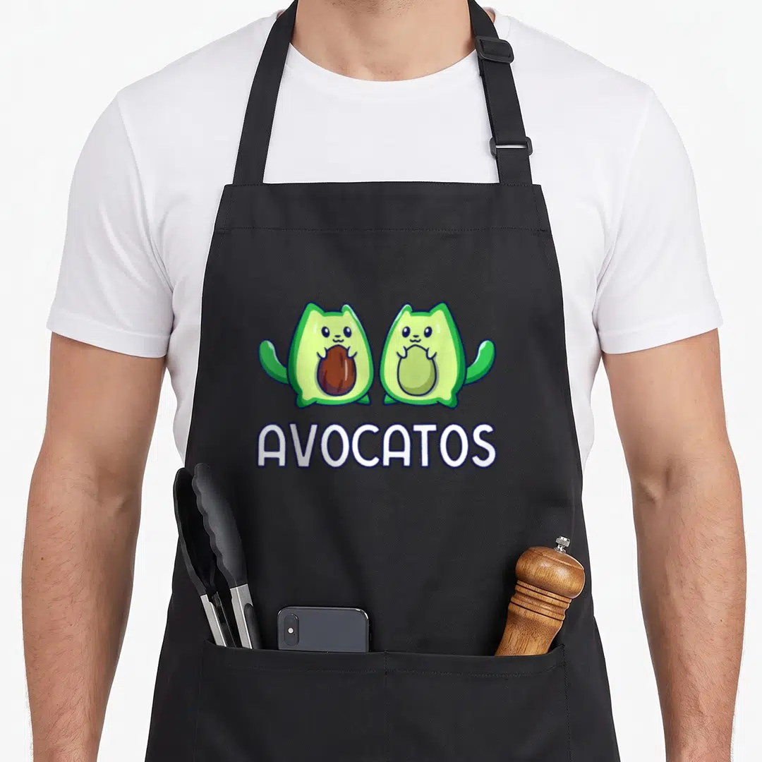 Cat avocado apron - Two cartoon avocado-cat hybrids with AVOCATOS text in green and white — shown on a black cotton canvas apron against a white background