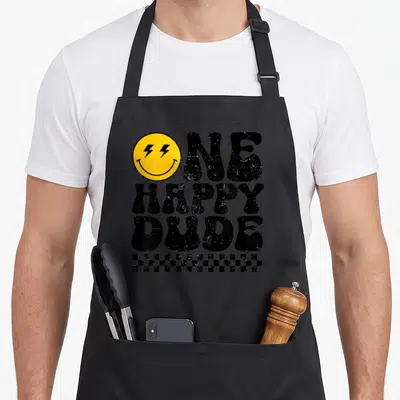 Dude custom face apron - Yellow smiley with lightning eyes and text ONE HAPPY DUDE above checkerboard strip — shown on a black cotton canvas apron against a white background
