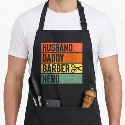Barber father day apron - Distressed orange yellow green grey bands with HUSBAND DADDY BARBER HERO text and scissors — shown on a black cotton canvas apron against a white background
