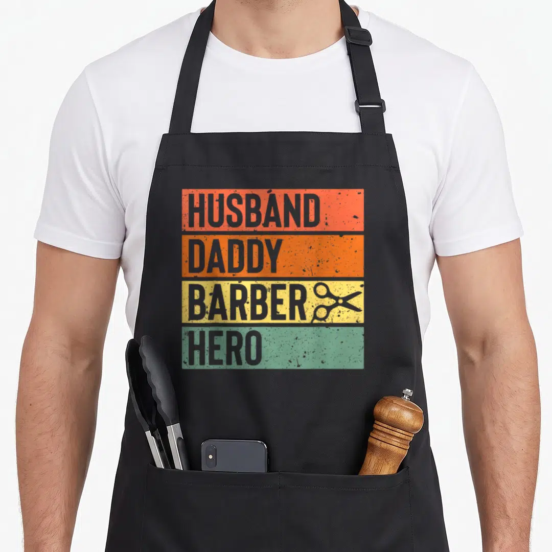Barber father day apron - Distressed orange yellow green grey bands with HUSBAND DADDY BARBER HERO text and scissors — shown on a black cotton canvas apron against a white background