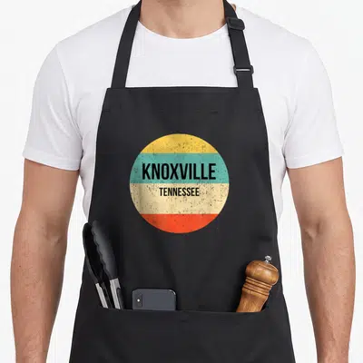 Funny tennessee knoxville apron - Circular retro badge with KNOXVILLE TENNESSEE text over yellow teal cream and red — shown on a black cotton canvas apron against a white background
