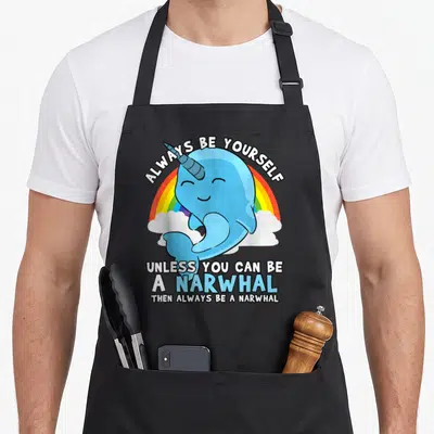 Narwhal unicorn apron - Blue cartoon narwhal with horn over rainbow and clouds with white text — shown on a black cotton canvas apron against a white background