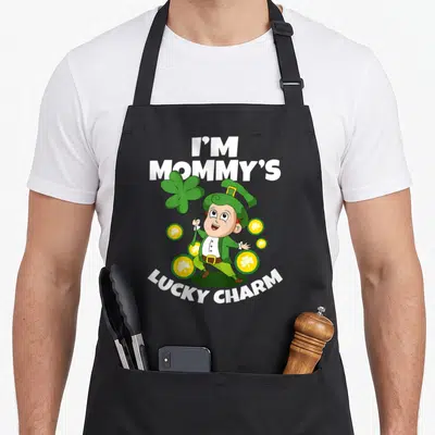 Charm toddler st patrick day aprons - Cartoon leprechaun holding green shamrock surrounded by yellow coins with I'M — shown on a black cotton canvas apron against a white background