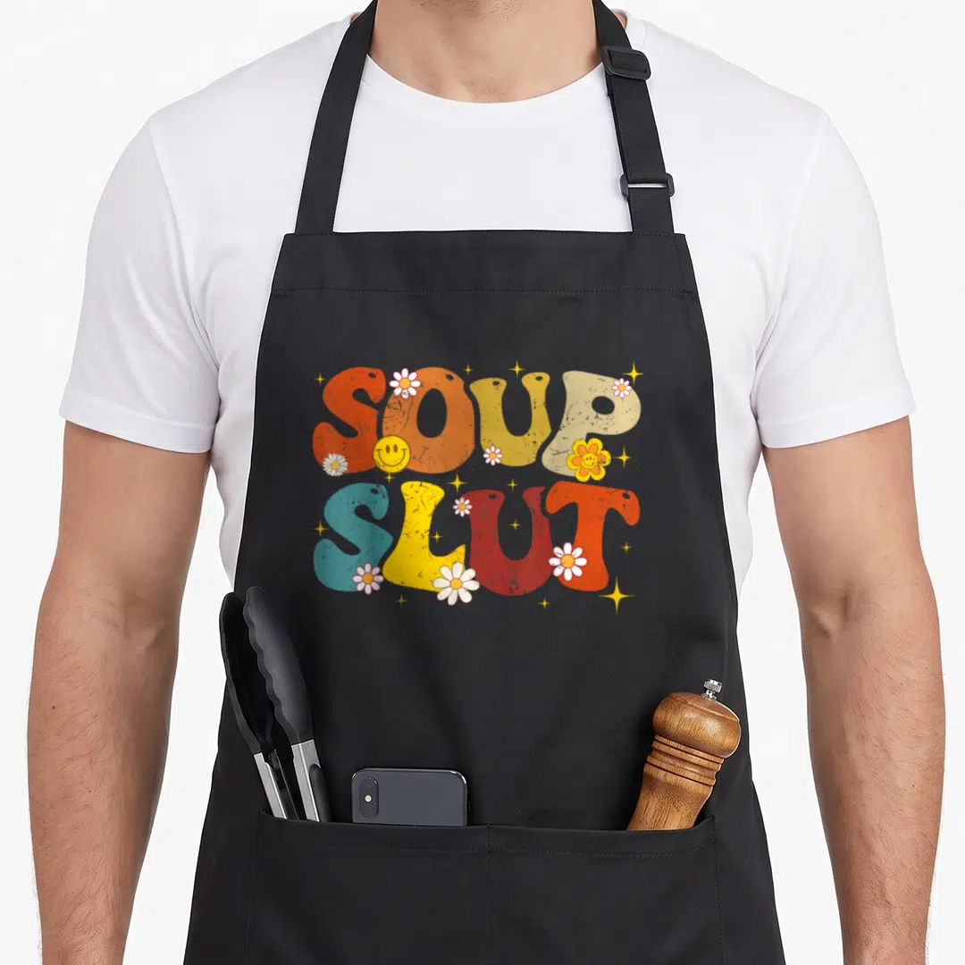 Soup christmas apron - Retro SOUP SLUT text with flowers, smiley face, and stars in orange yellow red blue beige — shown on a black cotton canvas apron against a white background