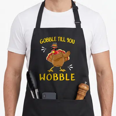 Gobble pilgrim apron - Cartoon turkey in sunglasses and pilgrim hat dabbing between yellow text — shown on a black cotton canvas apron against a white background