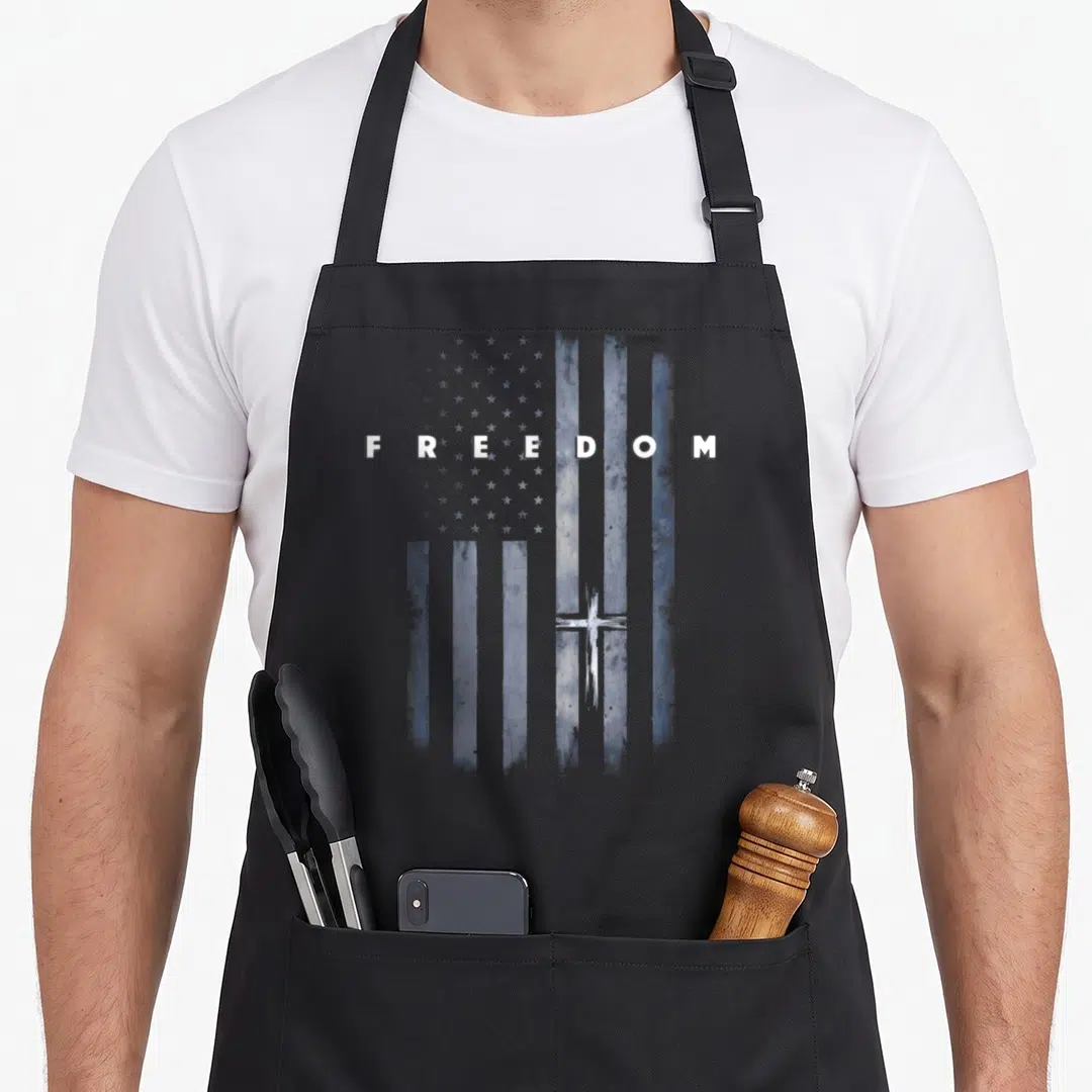 Grunge cross apron - Distressed blue and white USA flag with FREEDOM text and small cross in lower right — shown on a black cotton canvas apron against a white background