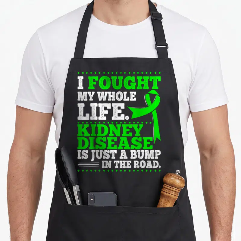 Ckd patient apron - Green ribbon and stacked text I fought my whole life kidney disease is just a bump in the road — shown on a black cotton canvas apron against a white background